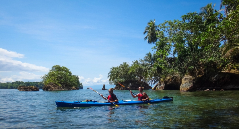 A two-person kayak is paddled on calm blue water around a small network of tree-covered islands.